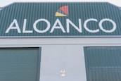 A large industrial building facade with the word 'ALOANCO' prominently displayed in white on a green background. Above the word, a multi-colored logo features a red, yellow, and orange triangular design. The building has green corrugated metal doors, and a small sign near the base indicates the presence of an alarm system.