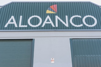 A large industrial building facade with the word 'ALOANCO' prominently displayed in white on a green background. Above the word, a multi-colored logo features a red, yellow, and orange triangular design. The building has green corrugated metal doors, and a small sign near the base indicates the presence of an alarm system.