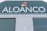 A large industrial building facade with the word 'ALOANCO' prominently displayed in white on a green background. Above the word, a multi-colored logo features a red, yellow, and orange triangular design. The building has green corrugated metal doors, and a small sign near the base indicates the presence of an alarm system.