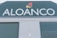 A large industrial building facade with the word 'ALOANCO' prominently displayed in white on a green background. Above the word, a multi-colored logo features a red, yellow, and orange triangular design. The building has green corrugated metal doors, and a small sign near the base indicates the presence of an alarm system.