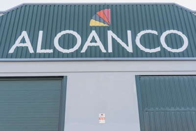 A large industrial building facade with the word 'ALOANCO' prominently displayed in white on a green background. Above the word, a multi-colored logo features a red, yellow, and orange triangular design. The building has green corrugated metal doors, and a small sign near the base indicates the presence of an alarm system.