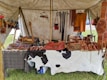 A market stall under a tent displays various handmade leather goods including sandals, bags, and straps. The items are organized neatly on a table covered with a cowhide. Brightly colored leather keychains or small crafts hang from a rod above. The scene suggests a rustic or traditional setting with a focus on artisanal craftsmanship.