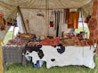 A market stall under a tent displays various handmade leather goods including sandals, bags, and straps. The items are organized neatly on a table covered with a cowhide. Brightly colored leather keychains or small crafts hang from a rod above. The scene suggests a rustic or traditional setting with a focus on artisanal craftsmanship.