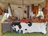 A market stall under a tent displays various handmade leather goods including sandals, bags, and straps. The items are organized neatly on a table covered with a cowhide. Brightly colored leather keychains or small crafts hang from a rod above. The scene suggests a rustic or traditional setting with a focus on artisanal craftsmanship.