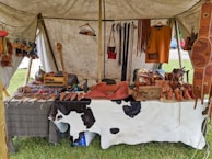 A market stall under a tent displays various handmade leather goods including sandals, bags, and straps. The items are organized neatly on a table covered with a cowhide. Brightly colored leather keychains or small crafts hang from a rod above. The scene suggests a rustic or traditional setting with a focus on artisanal craftsmanship.