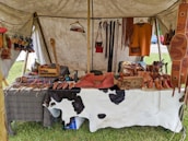 A market stall under a tent displays various handmade leather goods including sandals, bags, and straps. The items are organized neatly on a table covered with a cowhide. Brightly colored leather keychains or small crafts hang from a rod above. The scene suggests a rustic or traditional setting with a focus on artisanal craftsmanship.