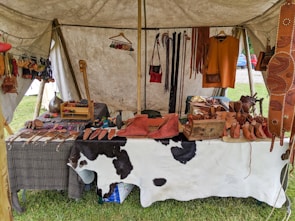 A market stall under a tent displays various handmade leather goods including sandals, bags, and straps. The items are organized neatly on a table covered with a cowhide. Brightly colored leather keychains or small crafts hang from a rod above. The scene suggests a rustic or traditional setting with a focus on artisanal craftsmanship.