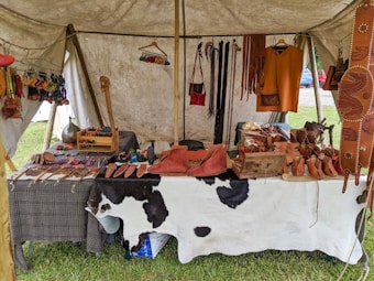 A market stall under a tent displays various handmade leather goods including sandals, bags, and straps. The items are organized neatly on a table covered with a cowhide. Brightly colored leather keychains or small crafts hang from a rod above. The scene suggests a rustic or traditional setting with a focus on artisanal craftsmanship.