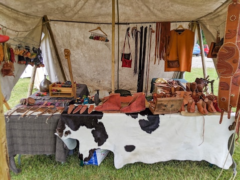 A market stall under a tent displays various handmade leather goods including sandals, bags, and straps. The items are organized neatly on a table covered with a cowhide. Brightly colored leather keychains or small crafts hang from a rod above. The scene suggests a rustic or traditional setting with a focus on artisanal craftsmanship.