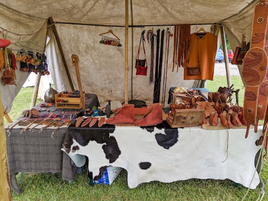 A market stall under a tent displays various handmade leather goods including sandals, bags, and straps. The items are organized neatly on a table covered with a cowhide. Brightly colored leather keychains or small crafts hang from a rod above. The scene suggests a rustic or traditional setting with a focus on artisanal craftsmanship.