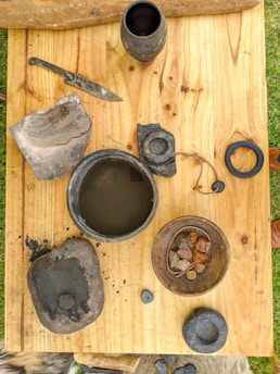 A wooden table displays various rustic items, including a knife, pottery, raw stones, and a leather cord. The pot contains a dark liquid, and a bowl holds several stones of varying colors.