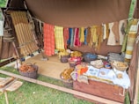 A rustic setup displaying a variety of yarns and woven textiles, with vibrant colors hanging from a tent. Basketfuls of colored yarns and wool are placed on the ground, and woven pieces are draped beside them. The ambiance suggests a traditional or artisanal craft setting.