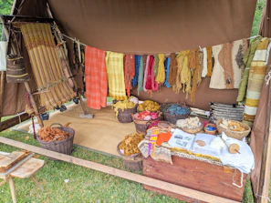 A rustic setup displaying a variety of yarns and woven textiles, with vibrant colors hanging from a tent. Basketfuls of colored yarns and wool are placed on the ground, and woven pieces are draped beside them. The ambiance suggests a traditional or artisanal craft setting.