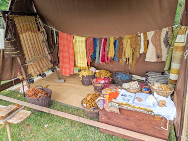 A rustic setup displaying a variety of yarns and woven textiles, with vibrant colors hanging from a tent. Basketfuls of colored yarns and wool are placed on the ground, and woven pieces are draped beside them. The ambiance suggests a traditional or artisanal craft setting.
