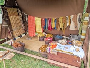 A rustic setup displaying a variety of yarns and woven textiles, with vibrant colors hanging from a tent. Basketfuls of colored yarns and wool are placed on the ground, and woven pieces are draped beside them. The ambiance suggests a traditional or artisanal craft setting.