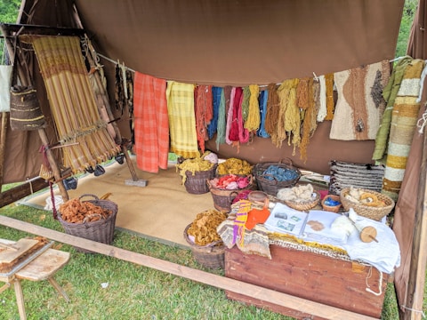 A rustic setup displaying a variety of yarns and woven textiles, with vibrant colors hanging from a tent. Basketfuls of colored yarns and wool are placed on the ground, and woven pieces are draped beside them. The ambiance suggests a traditional or artisanal craft setting.