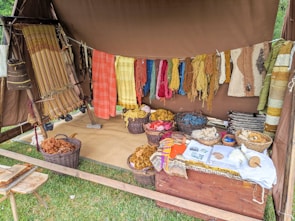 A rustic setup displaying a variety of yarns and woven textiles, with vibrant colors hanging from a tent. Basketfuls of colored yarns and wool are placed on the ground, and woven pieces are draped beside them. The ambiance suggests a traditional or artisanal craft setting.