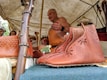A craftsman demonstrates leatherwork under a tent. On display are brown leather shoes with decorative patterns, hung on a wooden structure. Additional leather goods are visible in the background. Attendees appear interested as they observe.