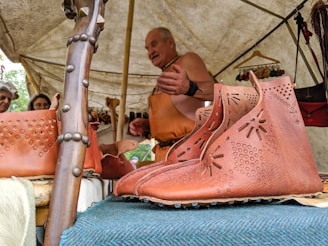 A craftsman demonstrates leatherwork under a tent. On display are brown leather shoes with decorative patterns, hung on a wooden structure. Additional leather goods are visible in the background. Attendees appear interested as they observe.