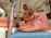 A craftsman demonstrates leatherwork under a tent. On display are brown leather shoes with decorative patterns, hung on a wooden structure. Additional leather goods are visible in the background. Attendees appear interested as they observe.
