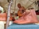 A craftsman demonstrates leatherwork under a tent. On display are brown leather shoes with decorative patterns, hung on a wooden structure. Additional leather goods are visible in the background. Attendees appear interested as they observe.