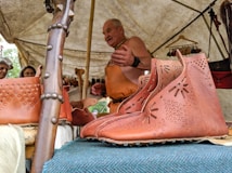 A craftsman demonstrates leatherwork under a tent. On display are brown leather shoes with decorative patterns, hung on a wooden structure. Additional leather goods are visible in the background. Attendees appear interested as they observe.