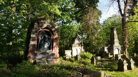 A serene cemetery scene surrounded by lush greenery and sunlight filtering through the trees. Several old tombstones and monuments are nestled among the foliage. Moss and plants are growing over some of the gravestones, adding to the tranquil and slightly mystical atmosphere.