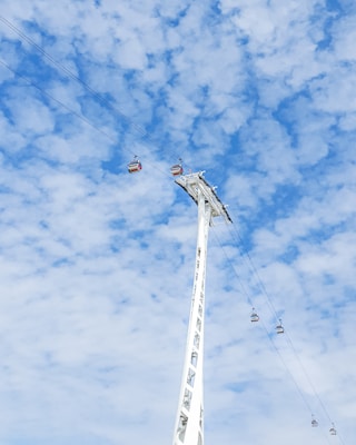 A tall, white cable car tower stretches upwards against a vast, blue sky filled with fluffy white clouds. Several cable cars are suspended from cables, moving across the sky.