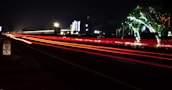 A nighttime scene with long exposure capturing red and white light trails of vehicles on a highway. There is festive lighting on trees, and a milestone indicates the distance to Chennai city. Bright lights from buildings are visible in the background.