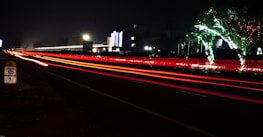 A nighttime scene with long exposure capturing red and white light trails of vehicles on a highway. There is festive lighting on trees, and a milestone indicates the distance to Chennai city. Bright lights from buildings are visible in the background.