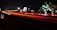A nighttime scene with long exposure capturing red and white light trails of vehicles on a highway. There is festive lighting on trees, and a milestone indicates the distance to Chennai city. Bright lights from buildings are visible in the background.