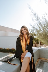 A person wearing a graduation gown and sash sits on a concrete ledge outdoors. The setting appears to be a campus with modern buildings and landscaping visible in the background. The individual has long hair and smiles at the camera. A graduation cap with a tassel lies beside them on the ledge.
