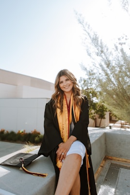 A person wearing a graduation gown and sash sits on a concrete ledge outdoors. The setting appears to be a campus with modern buildings and landscaping visible in the background. The individual has long hair and smiles at the camera. A graduation cap with a tassel lies beside them on the ledge.