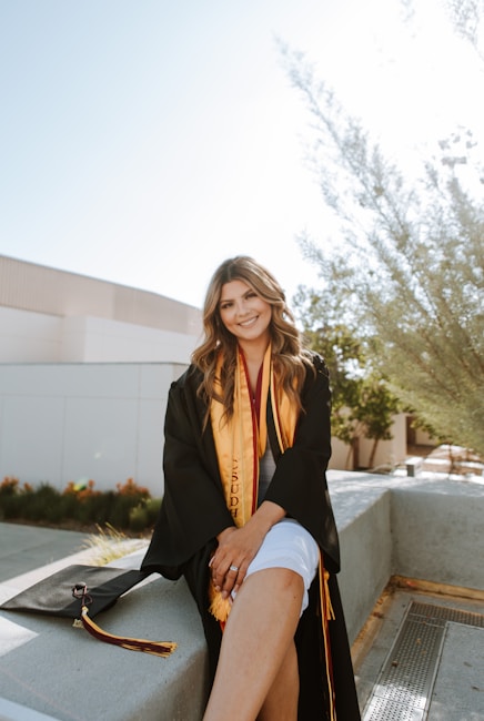 A person wearing a graduation gown and sash sits on a concrete ledge outdoors. The setting appears to be a campus with modern buildings and landscaping visible in the background. The individual has long hair and smiles at the camera. A graduation cap with a tassel lies beside them on the ledge.
