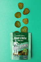 A packet of SuckerPunch Bread & Butter Pickle Chips is laid flat on a vibrant green surface. Several sliced pickle chips are arranged in an irregular pattern above the packet. The packaging prominently features a vintage-style illustration of a man in boxing attire, and includes labels emphasizing the product's snappy sweet flavor and non-GMO status.