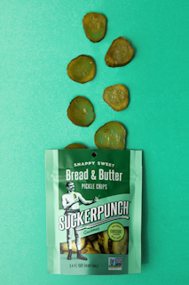 A packet of SuckerPunch Bread & Butter Pickle Chips is laid flat on a vibrant green surface. Several sliced pickle chips are arranged in an irregular pattern above the packet. The packaging prominently features a vintage-style illustration of a man in boxing attire, and includes labels emphasizing the product's snappy sweet flavor and non-GMO status.