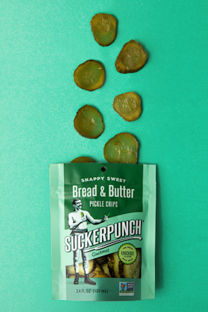 A packet of SuckerPunch Bread & Butter Pickle Chips is laid flat on a vibrant green surface. Several sliced pickle chips are arranged in an irregular pattern above the packet. The packaging prominently features a vintage-style illustration of a man in boxing attire, and includes labels emphasizing the product's snappy sweet flavor and non-GMO status.