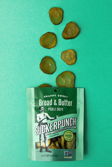 A packet of SuckerPunch Bread & Butter Pickle Chips is laid flat on a vibrant green surface. Several sliced pickle chips are arranged in an irregular pattern above the packet. The packaging prominently features a vintage-style illustration of a man in boxing attire, and includes labels emphasizing the product's snappy sweet flavor and non-GMO status.