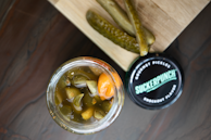 A jar filled with pickled vegetables, including sliced pickles and a noticeable orange pepper, placed next to a wooden board with sliced pickles on top. The jar lid nearby features the brand 'SuckerPunch' labeled as gourmet pickles with a knockout flavor.