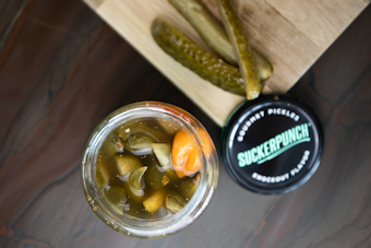 A jar filled with pickled vegetables, including sliced pickles and a noticeable orange pepper, placed next to a wooden board with sliced pickles on top. The jar lid nearby features the brand 'SuckerPunch' labeled as gourmet pickles with a knockout flavor.