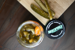 A jar filled with pickled vegetables, including sliced pickles and a noticeable orange pepper, placed next to a wooden board with sliced pickles on top. The jar lid nearby features the brand 'SuckerPunch' labeled as gourmet pickles with a knockout flavor.