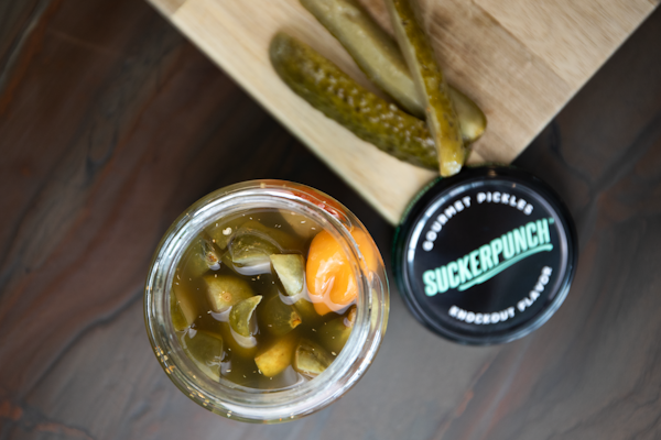A jar filled with pickled vegetables, including sliced pickles and a noticeable orange pepper, placed next to a wooden board with sliced pickles on top. The jar lid nearby features the brand 'SuckerPunch' labeled as gourmet pickles with a knockout flavor.