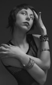 A grayscale portrait of a person with short hair, tilting their head and touching their forehead with one hand while the other arm is crossed over the torso. They are wearing a mesh top and several bracelets, the image exudes a sense of elegance and introspection.