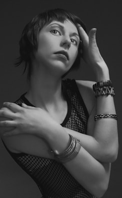 A grayscale portrait of a person with short hair, tilting their head and touching their forehead with one hand while the other arm is crossed over the torso. They are wearing a mesh top and several bracelets, the image exudes a sense of elegance and introspection.