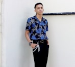woman in blue and white floral button up shirt and black pants standing beside white wall