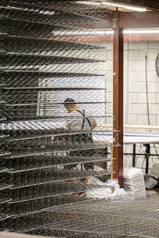 A person wearing a dark apron stands in an industrial setting surrounded by stacks of metal spring frames. The frames are organized in layers, creating a repetitive pattern. The environment includes metal structures and a workspace equipped with tools and materials. Lighting is provided by overhead fluorescent lights, giving the scene a functional atmosphere.