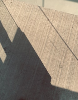 Shadows of linear objects cast on a textured concrete surface. The lines of the shadows create geometric patterns, and the texture of the concrete adds depth to the image.