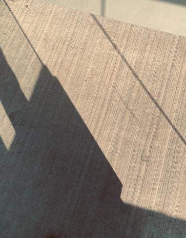 Shadows of linear objects cast on a textured concrete surface. The lines of the shadows create geometric patterns, and the texture of the concrete adds depth to the image.