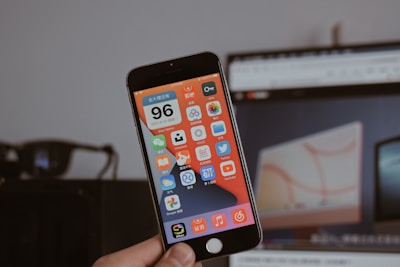 A person is holding a smartphone displaying a screen filled with colorful app icons, mostly in Chinese language. The background is blurred, featuring a computer monitor showing a webpage and a dark object resembling glasses.