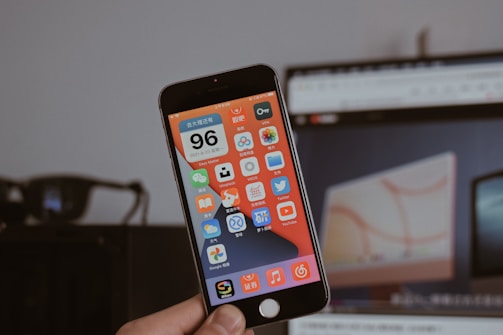 A person is holding a smartphone displaying a screen filled with colorful app icons, mostly in Chinese language. The background is blurred, featuring a computer monitor showing a webpage and a dark object resembling glasses.